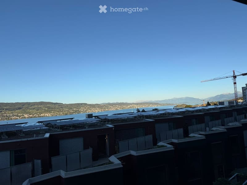 Roof terrace with solar panels, modern buildings, scenic view of water and distant mountains.