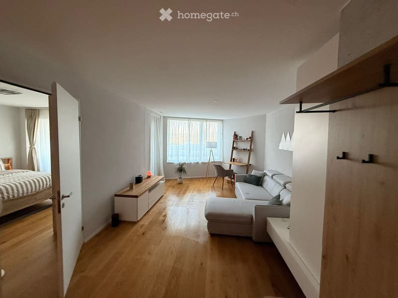 A modern, open-concept living and bedroom space with light wooden flooring, a grey sectional sofa, a bed with striped bedding, a small desk with a chair, and a shelf with decorative items.