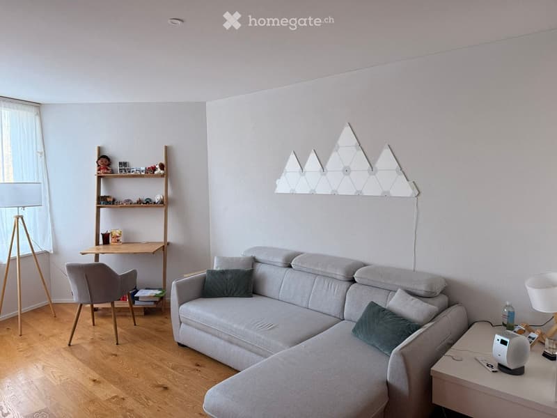 Modern living room with a grey sectional sofa, wooden floor, a small wooden desk, a shelf with decorative items, and a geometric mirror on the wall.