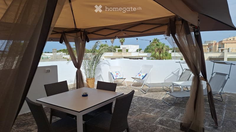 A sunny outdoor terrace with a large umbrella, a dining table with chairs, lounge chairs, and a potted plant, overlooking a cityscape with palm trees.