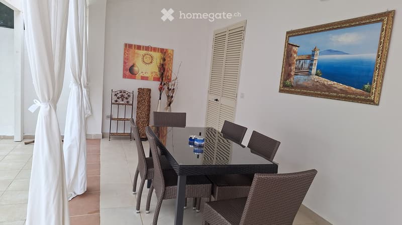 A modern dining area with a black glass table, wicker chairs, a vase with flowers, a shelf, and two paintings on the walls.