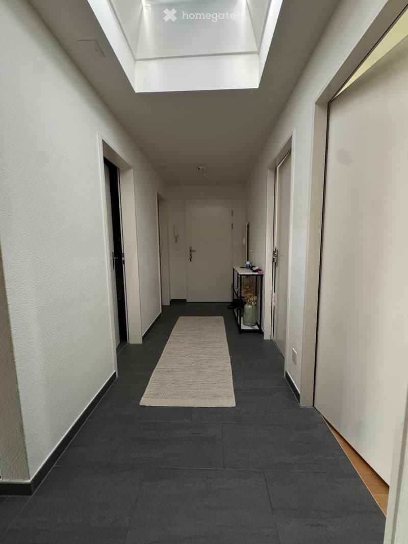 Clean, modern hallway with dark tiled flooring, white walls, and a small beige rug in the center. A small table with a vase is placed against the right wall.