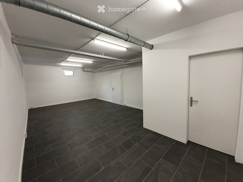 Empty room with white walls, dark tiled floor, and exposed pipes on the ceiling. No furniture or equipment.