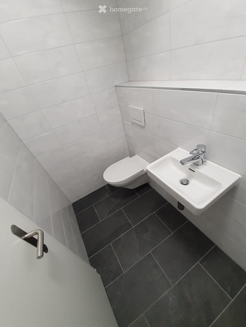 Modern bathroom with white tiled walls, dark grey tiled floor, toilet, and a white sink with a chrome faucet.