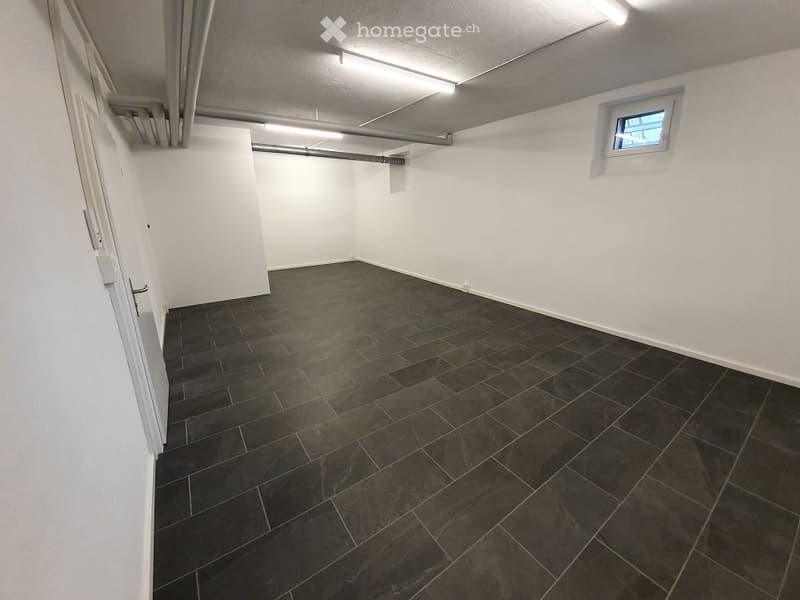 Empty room with white walls, dark tiled floor, and a small window. No furniture or equipment present.