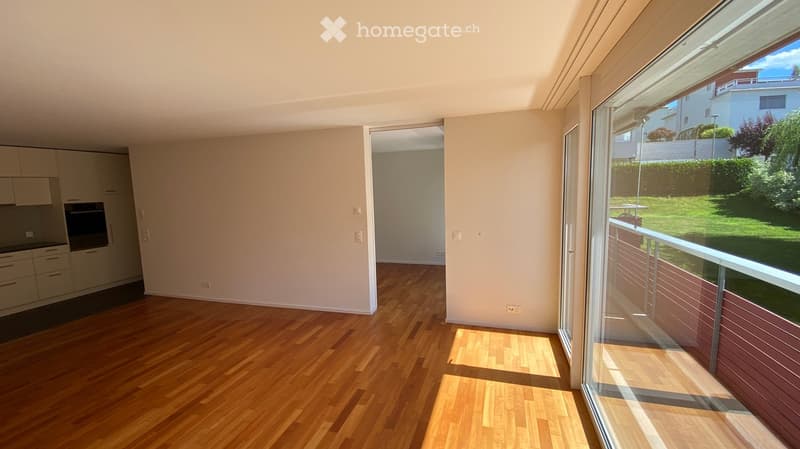 Empty room with wooden flooring, white walls, and large windows offering a view of a green garden. The room is unfurnished and has a modern, minimalist design.