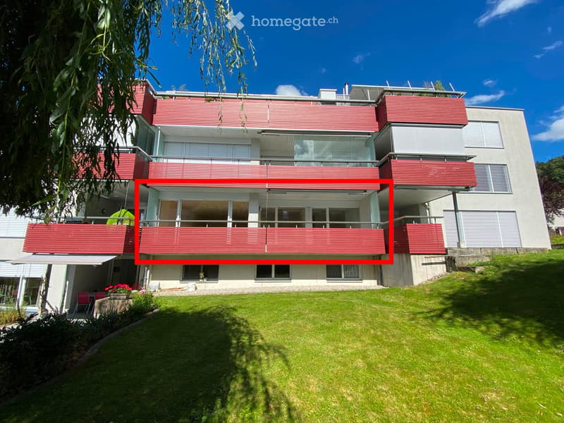 Modern multi-story building with red and white exterior, large windows, balconies, and a well-maintained lawn in front.