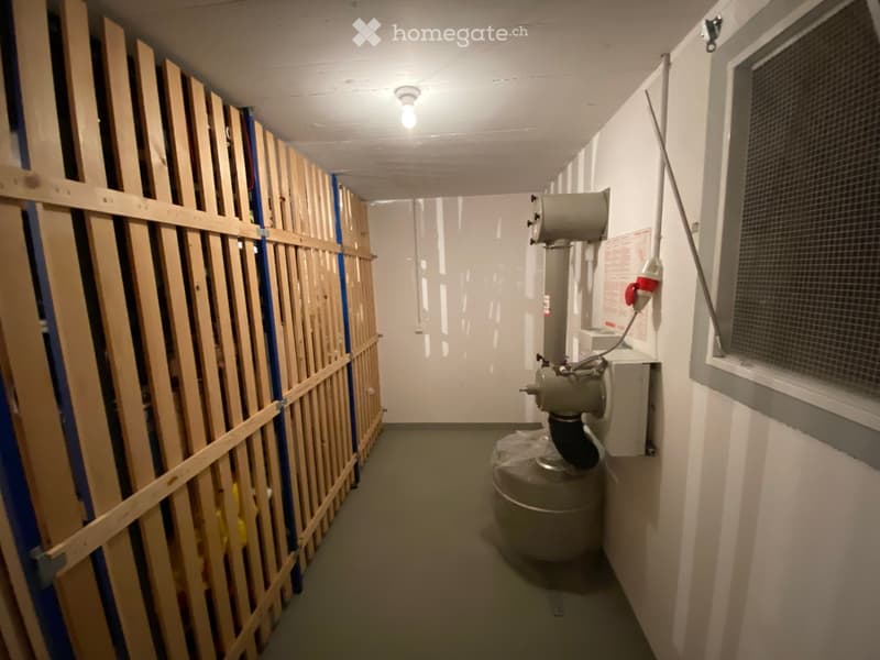 A narrow hallway under construction with exposed wooden framing on one side, a ventilation unit on the opposite wall, and a small window at the end.