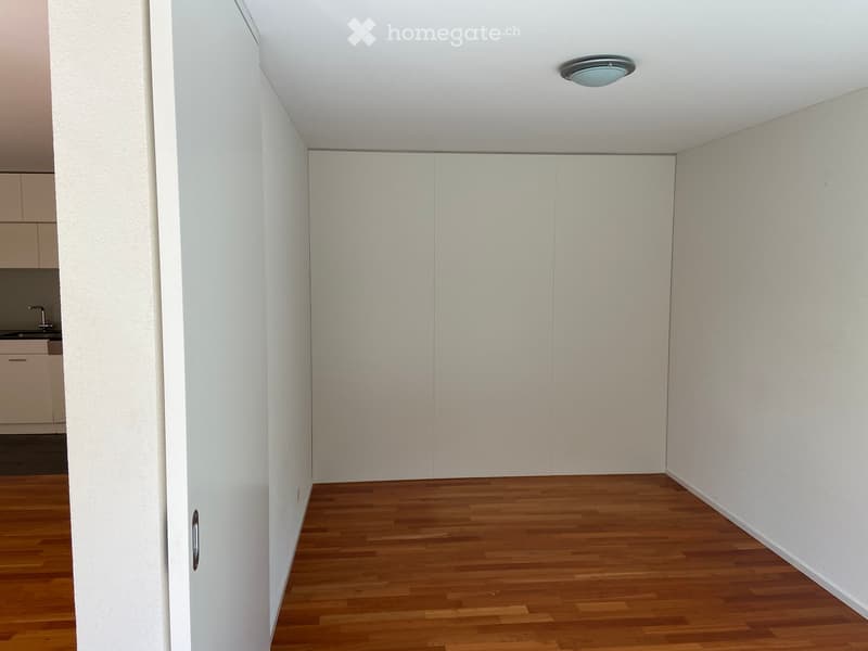 Empty room with wooden flooring, white walls, and a closed white closet door. A small section of a kitchen with cabinets and a sink is visible in the background.