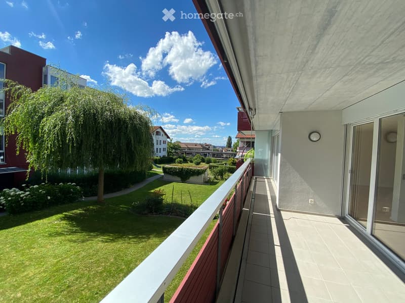 Balcony with tiled floor, wooden railing, and view of a well-maintained garden with a large weeping tree, lawn, and surrounding buildings.