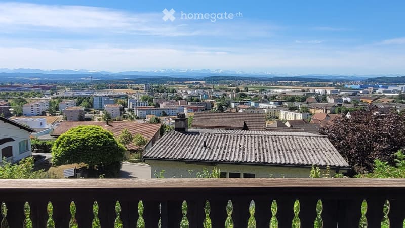 A balcony overlooking a town with houses, trees, and distant mountains under a clear blue sky.
