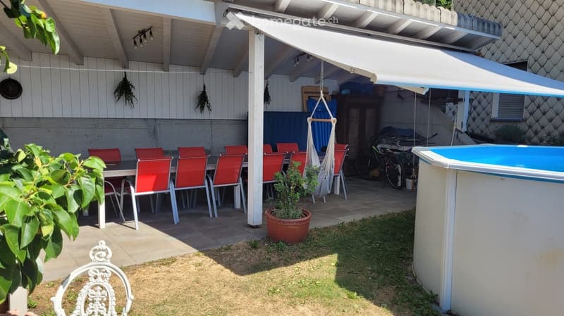 Outdoor patio area with a dining table and red chairs, a small swimming pool, potted plants, and a covered seating area with hanging decorations.