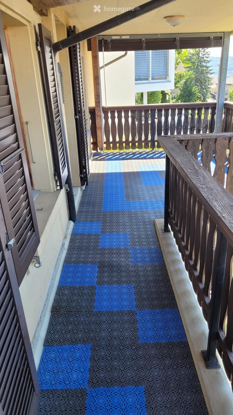Balcony with wooden railing, patterned blue mats on the floor, and a view of the surrounding area.