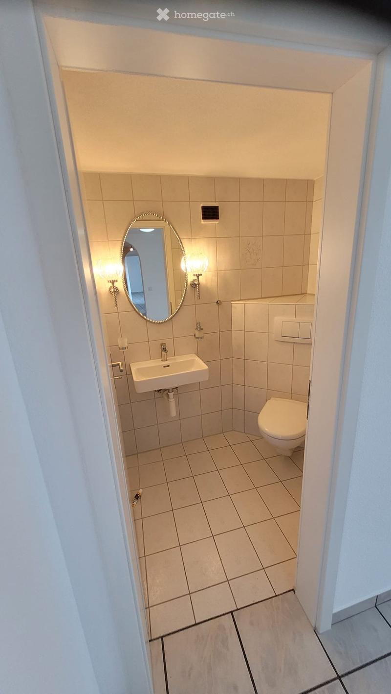 Small bathroom with beige tiled walls and floor, featuring a sink, toilet, and a round mirror with a decorative frame.