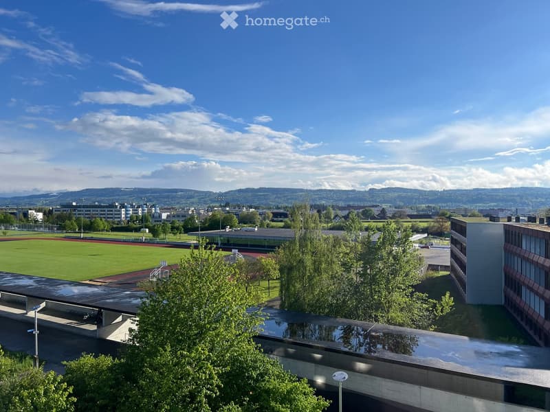 View of a sports field with a running track, surrounded by trees and buildings, under a clear blue sky with scattered clouds.