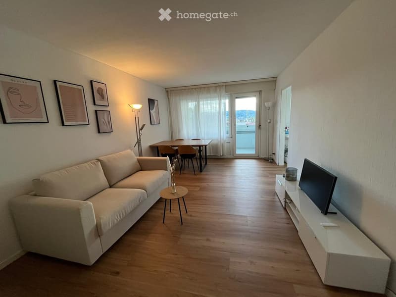 Bright living room with wooden flooring, a beige sofa, a small coffee table, a dining area with a table and chairs, a TV stand, and a floor lamp. Large windows and a balcony are visible.