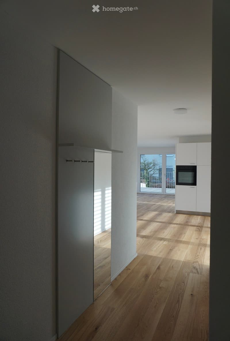 A modern, minimalist hallway with light wooden flooring, white walls, and a recessed ceiling light. The hallway leads to a kitchen area with white cabinets and a built-in oven, visible through an open doorway.