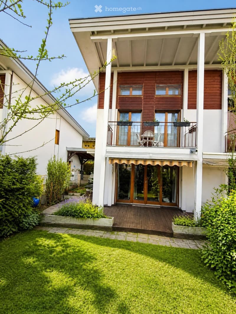 Two-story house with a wooden balcony, large windows, and a well-maintained garden with a wooden deck and patio furniture.