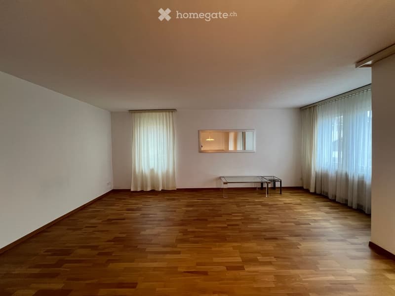 Empty room with wooden flooring, large windows with curtains, and a small glass table with a mirror on the wall.