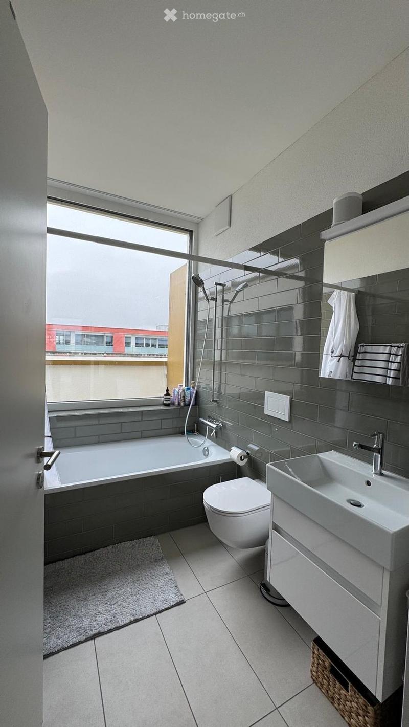 Modern bathroom with a bathtub, toilet, sink, and showerhead. Features dark grey tiles, a large window, and a white towel on the mirror.