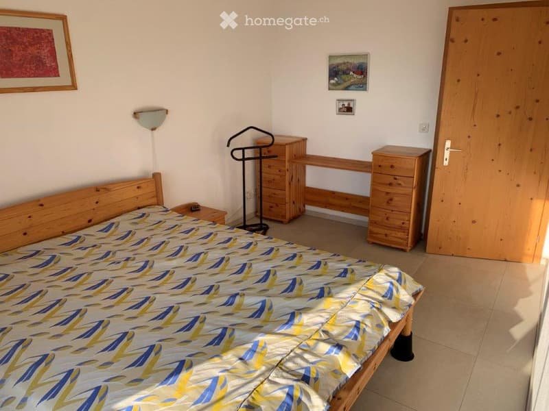Cozy bedroom with a wooden bed, nightstand, wardrobe, coat rack, and wall-mounted lamp. The bed has a blue and yellow patterned duvet cover.