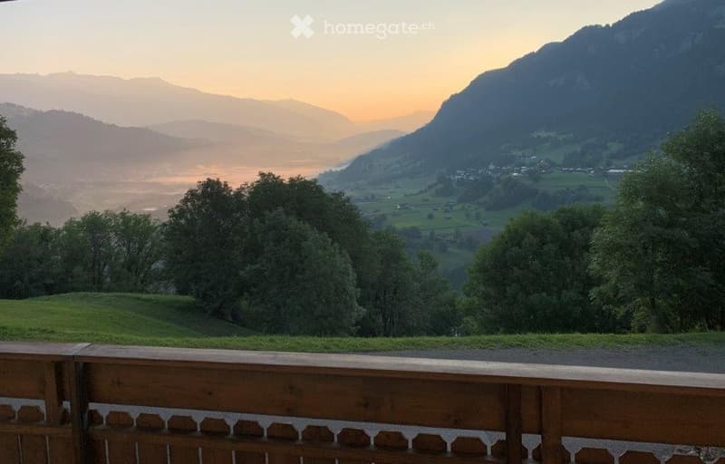 Balcony with wooden railing overlooking a scenic valley and mountains at sunset.