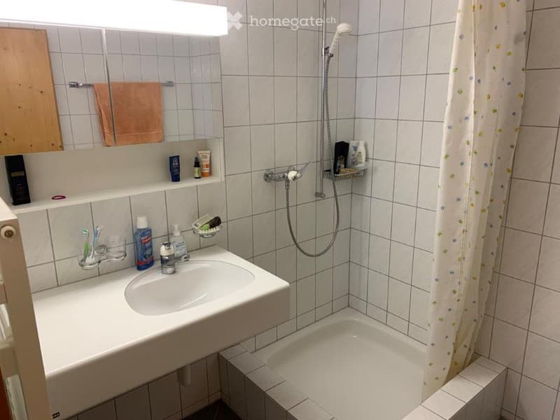 A compact bathroom with a white tiled wall, a sink with a mirror above it, a shower area with a curtain, and a small cabinet with toiletries.