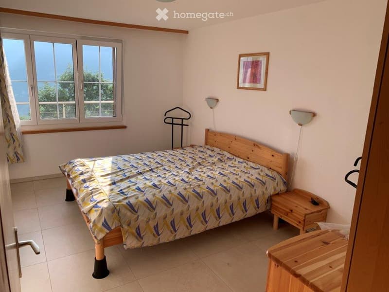 Cozy bedroom with a wooden bed, colorful bedspread, nightstand, wall-mounted lamp, and a view through large windows.