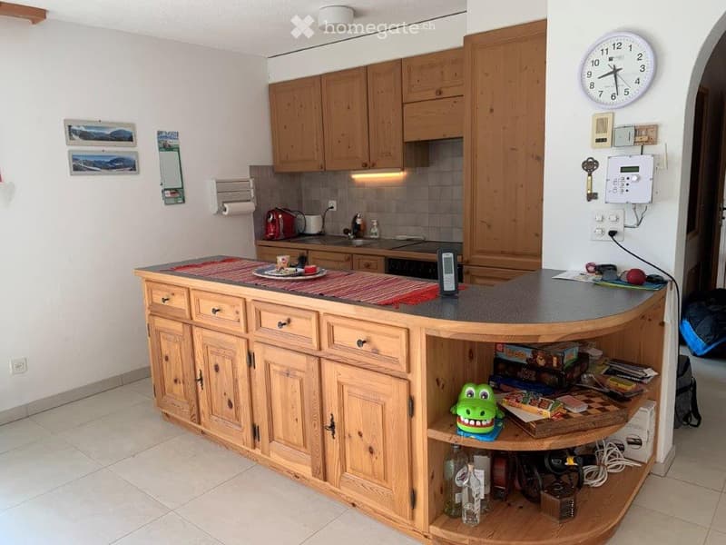 A well-lit kitchen with wooden cabinets, a central island counter, and various appliances including a toaster, fridge, and oven. The counter has a red placemat and a toy frog on the lower shelf.