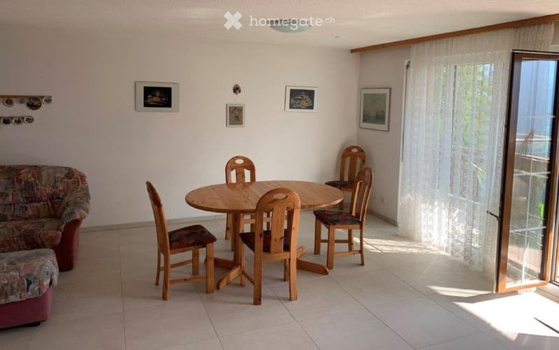 A cozy living and dining area with a patterned sofa, a round wooden dining table, and six wooden chairs. The room features light-colored walls, framed artwork, and a large glass door leading to a balcony.