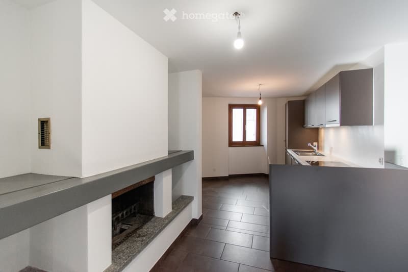 An unfurnished room with a fireplace, tiled floor, and a window. The space includes a kitchen area with cabinets and a sink, but no furniture or appliances are present.