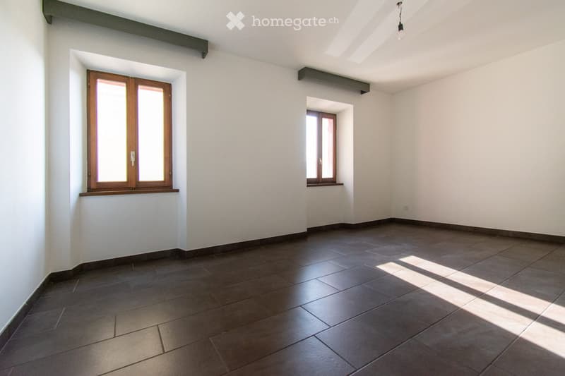 Empty room with dark tiled floor, white walls, and two windows with wooden frames. Natural light streams in from the windows, casting shadows on the floor.