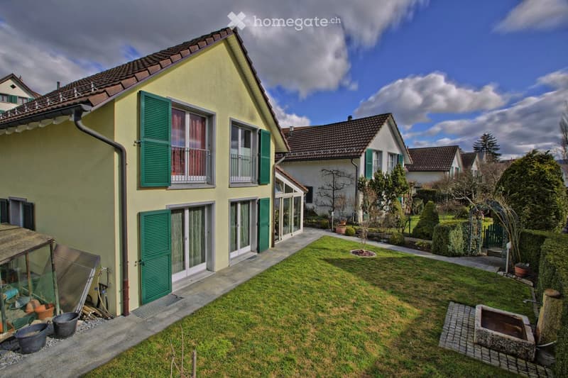 Two-story house with yellow exterior, green shutters, and multiple windows. The house features a well-maintained lawn, a small garden area with potted plants, and a paved pathway leading to the entrance.