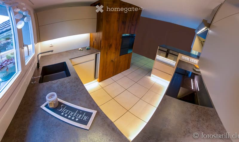 Modern kitchen with light wood cabinets, stainless steel sink, and a central island countertop. Features a candle and a decorative sign on the island.