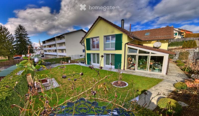 A two-story yellow house with green shutters, a well-maintained garden, and a small greenhouse attached to the side.