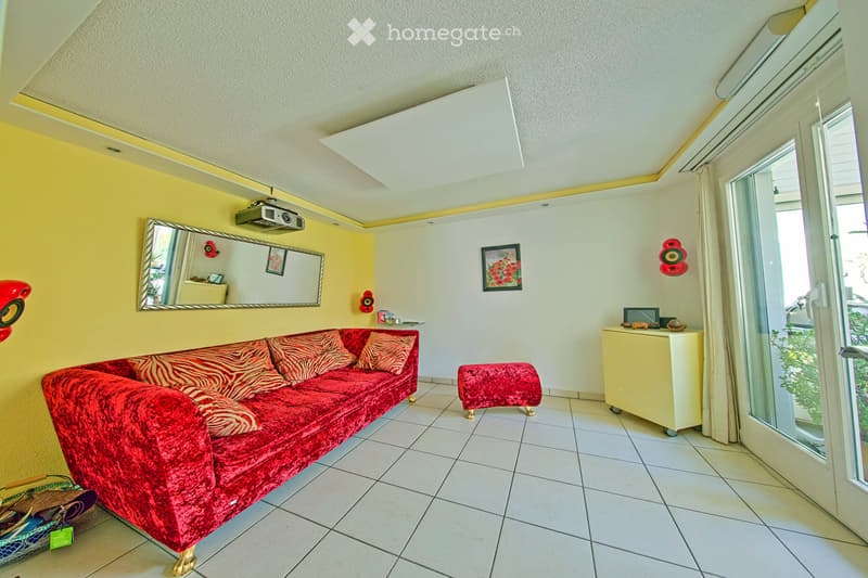 Bright living room with red velvet sofa, matching ottoman, small table, mirror, projector, and picture frames on the walls.