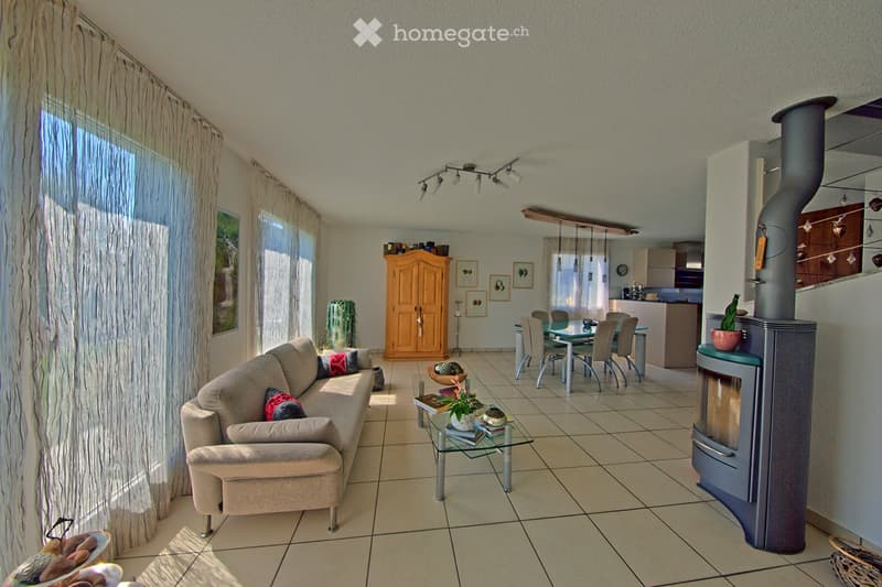 Bright living room with a beige sofa, glass coffee table, fireplace, dining area with a table and chairs, and a kitchen area in the background.