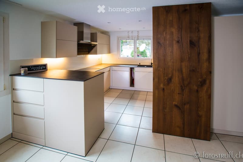 Modern kitchen with white cabinets, a central island, and a large window. Features include a range hood, sink, and a wooden panel accent wall.