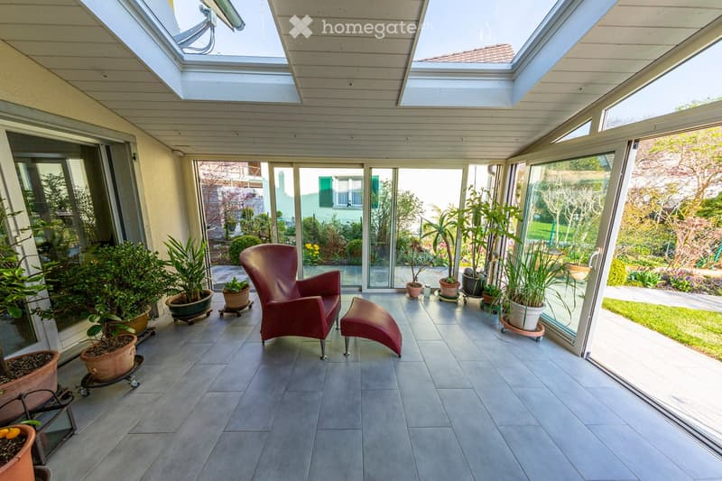 Bright sunroom with large glass walls and skylights, featuring a red leather chair, matching stool, and several potted plants arranged around the space.