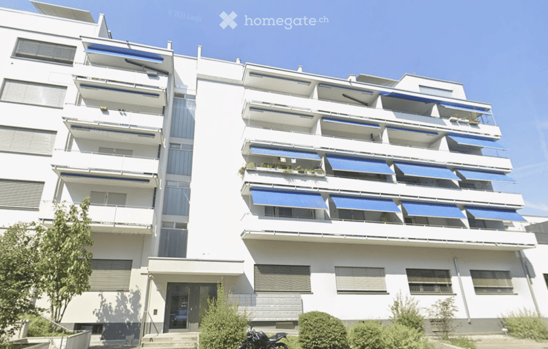 Modern multi-story residential building with white facade, balconies, blue awnings, and small garden area in front.