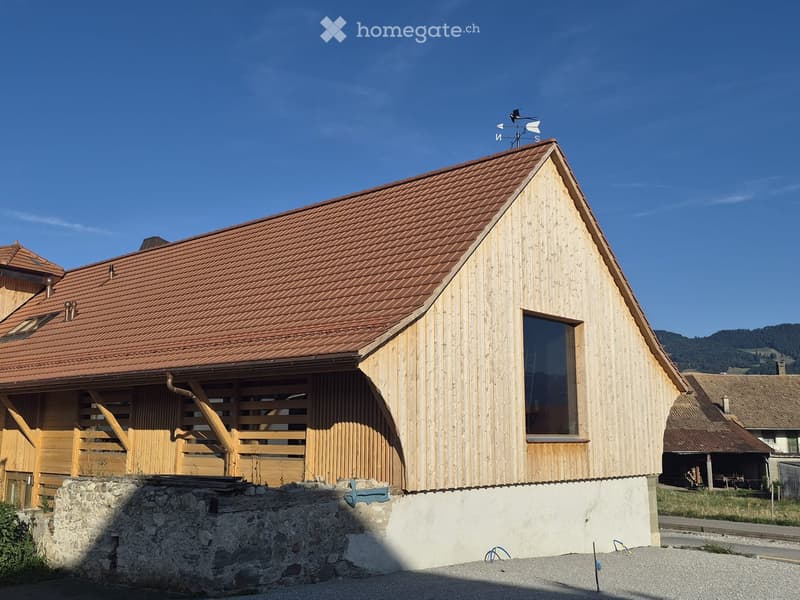 A modern wooden house with a red-tiled roof, large windows, and a stone base, featuring a weather vane on the roof.