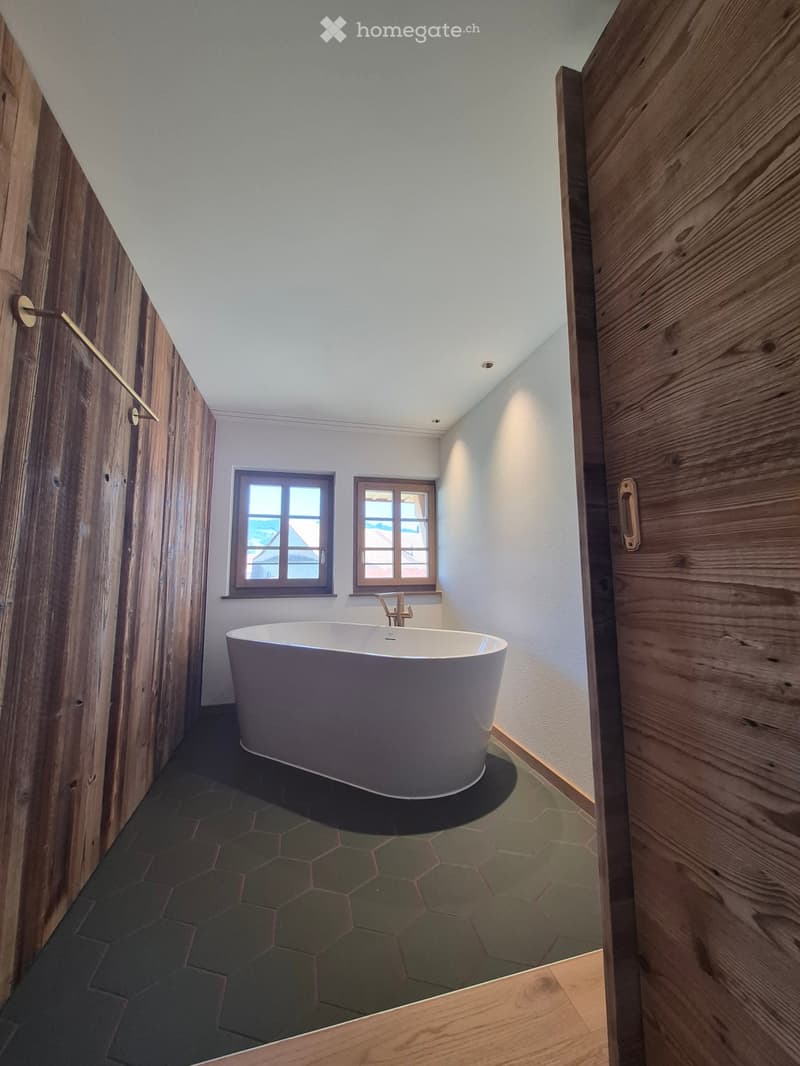 Modern bathroom with a freestanding bathtub, wooden walls, and two windows offering a view.