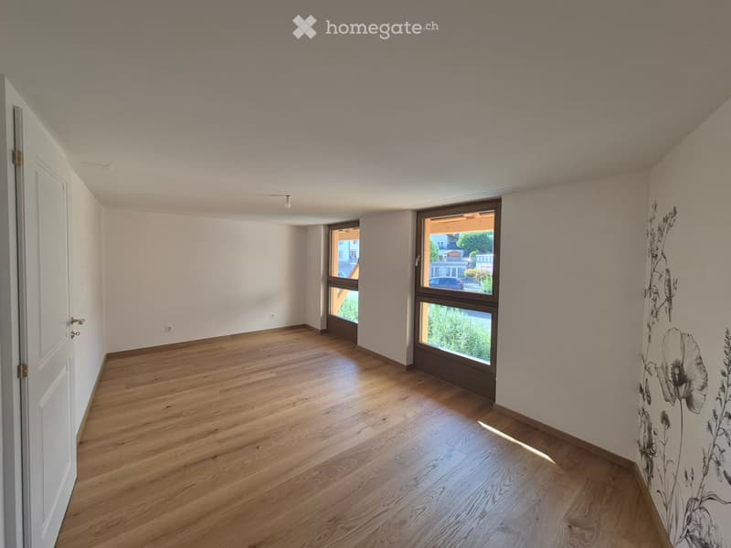 Empty room with wooden flooring, white walls, and large windows offering a view of the outside. The room has a door on the left and a wall with a floral design on the right.
