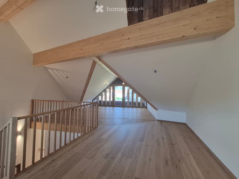 Spacious, open hallway with wooden flooring and exposed wooden beams. Features a wooden staircase with a railing leading to an upper level, and large windows at the end allowing natural light to flood the space.