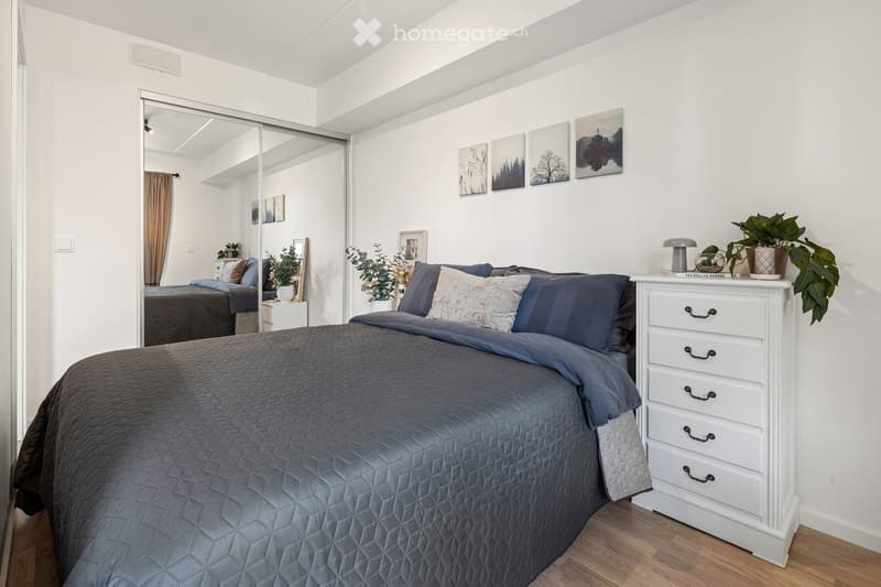 Modern bedroom with a large bed, nightstand, mirror, and decorative plants. The room features white walls, wooden flooring, and artwork on the walls.