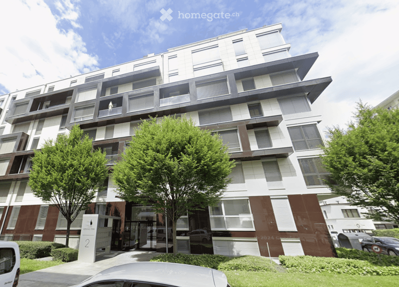 Modern multi-story residential building with balconies, large windows, and contemporary design. The building features a mix of white and brown tones, with greenery in the foreground and a clear blue sky above.
