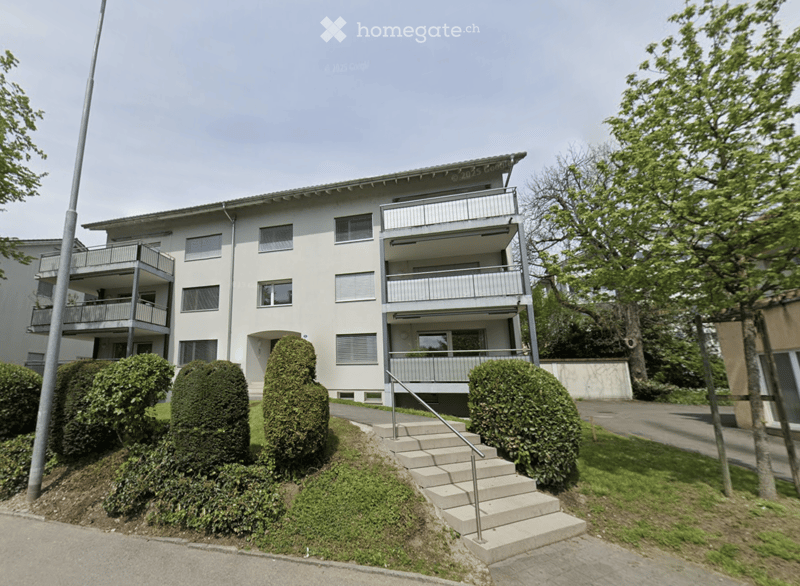 A modern, multi-story residential building with balconies and a raised ground floor entrance, surrounded by trimmed hedges and trees.
