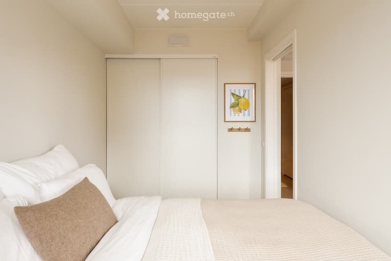 A modern bedroom with a neatly made bed, white bedding, a brown throw pillow, a built-in wardrobe, and a framed picture of lemons on the wall.