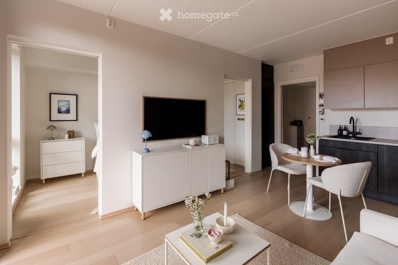 Modern, open-concept living and dining area with light wood flooring, white walls, and contemporary furniture including a sofa, coffee table, dining table with chairs, and a kitchen area with a sink and cabinets.