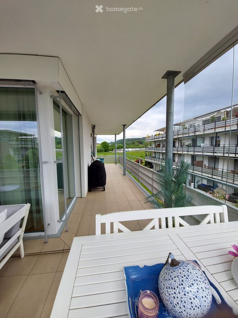 Balcony with a white table, chairs, a decorative vase, and a candle, overlooking a residential area with other apartment buildings and greenery.